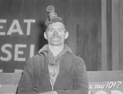 A man with coal dust on his face and a miner's cap lamp on his head, arms folded, seated in front of a painted wooden sign.