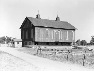 A large two-story wooden barn with tall arched louvered windows and two cupolas on its roof, with a small shed, a dirt lane and a rail fence in front.
