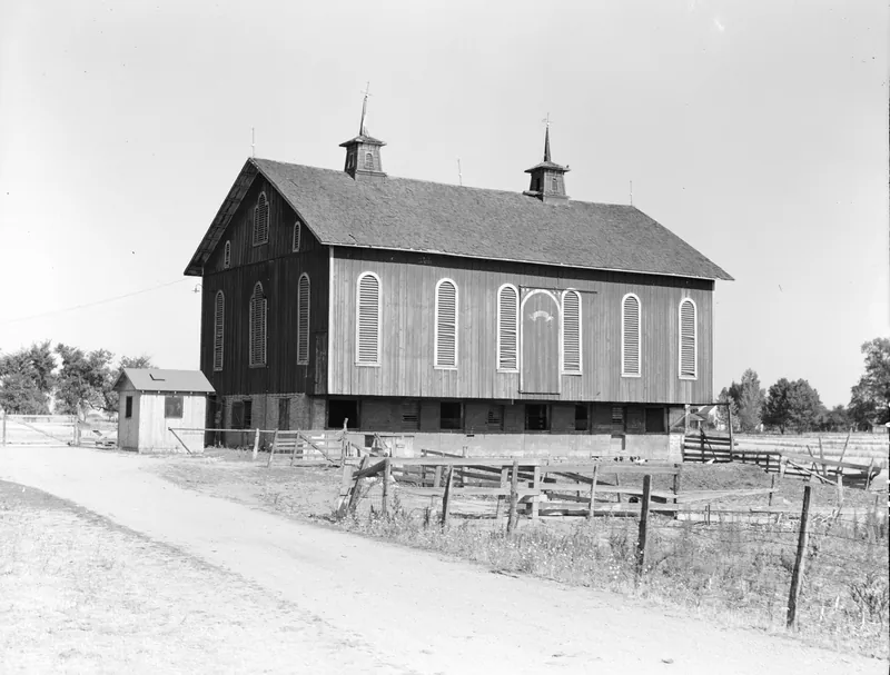 A large two-story wooden barn with tall arched louvered windows and two cupolas on its roof, with a small shed, a dirt lane and a rail fence in front.