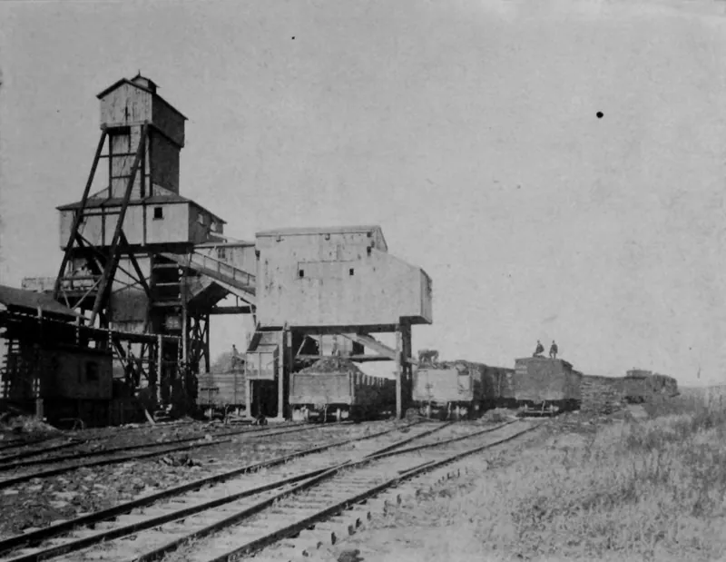 A tall wooden headframe and tipple over several railroad tracks, with a line of coal cars and two men standing on top of a car.