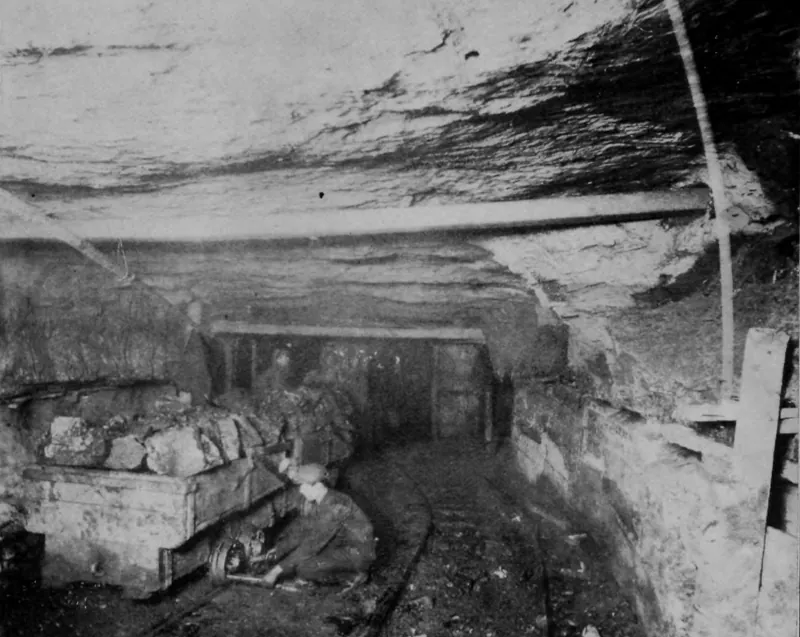 An underground passage with a low rock roof braced by timbers; a miner crouches beside a mine car heaped with lumps of coal on narrow rails.