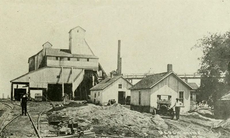 A large wooden tipple building with a tall peaked top stands beside railroad tracks. Two small frame buildings, an early automobile and two men are in front, with a smokestack and an overhead conveyor behind.