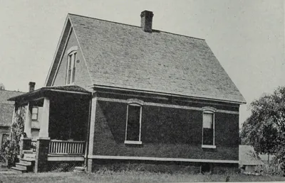 A one-and-a-half-story brick house with a steep shingled roof and a front porch, on a grassy lot.