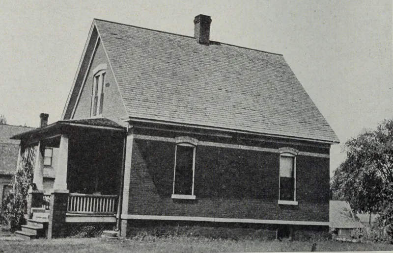 A one-and-a-half-story brick house with a steep shingled roof and a front porch, on a grassy lot.