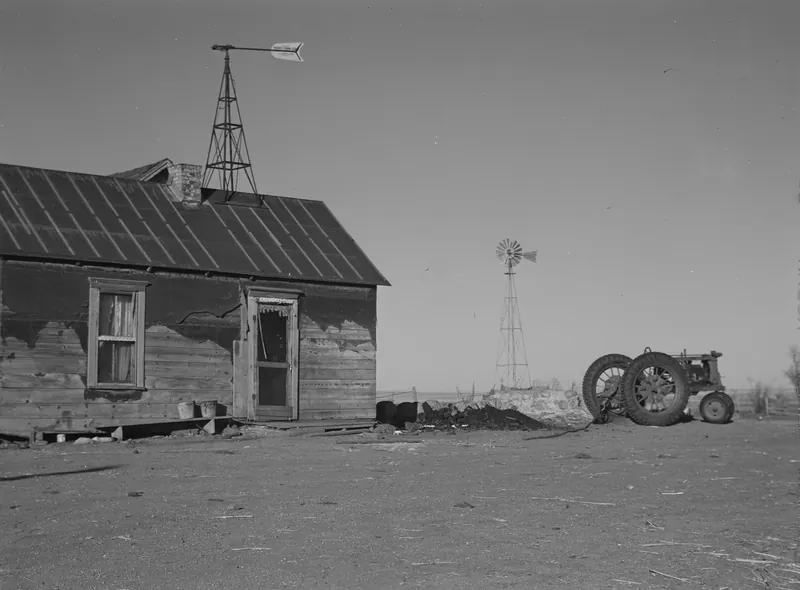 A weathered wooden farmhouse with a wind vane tower on its roof, a windmill and a tractor beside it, on flat open prairie.