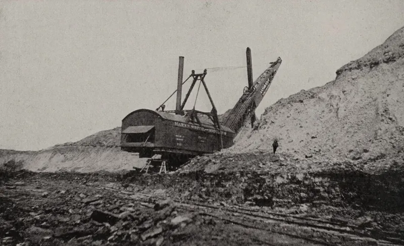 A large steam stripping shovel with two smokestacks and a long boom digging into a high bank of earth, with a worker standing on the slope and rails in the broken ground below.