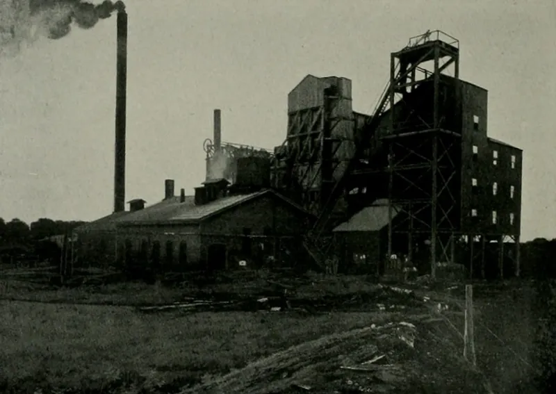 A large coal mine plant: a tall steel headframe and multi-story tipple, a brick engine house, and a smokestack trailing black smoke over a rough field.