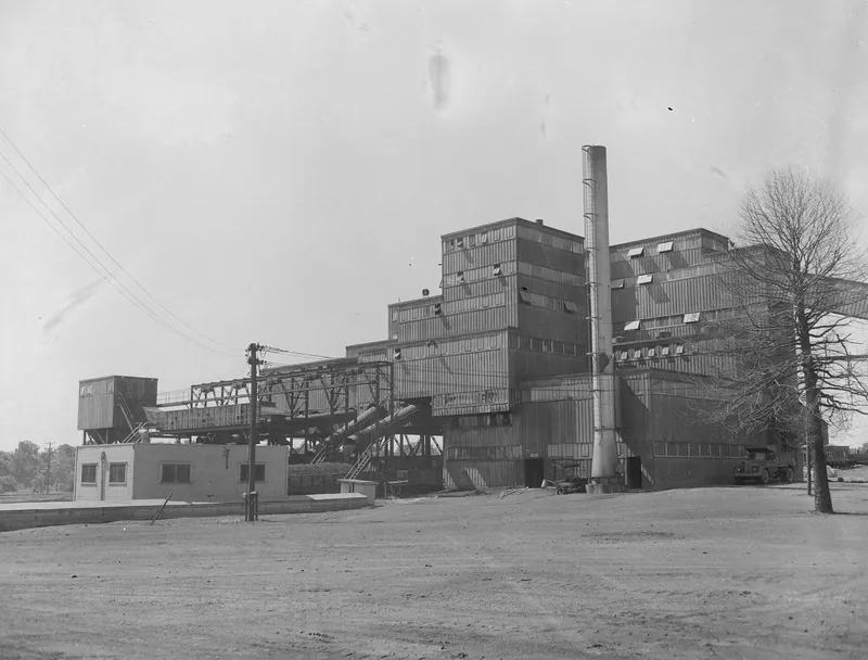 A large multi-story corrugated metal coal tipple and preparation plant with a tall smokestack and conveyor bridges, seen across a bare dirt yard with a small office building and a bare tree.