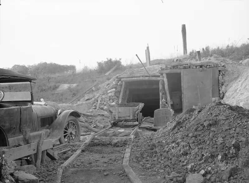 A timber-framed mine entrance cut into a hillside, with a wooden mine car on narrow rails in front, a heap of coal at right and an old automobile at left.