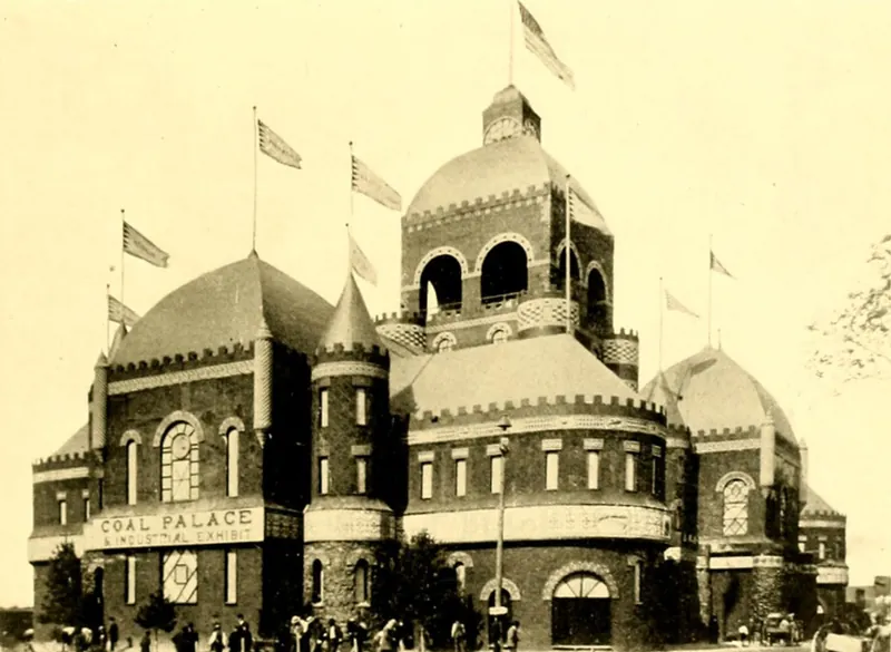 A large castle-like hall with a central domed tower, round corner towers and flags, its walls built of blocks of coal, with a sign reading Coal Palace and Industrial Exhibit and people gathered at the base.