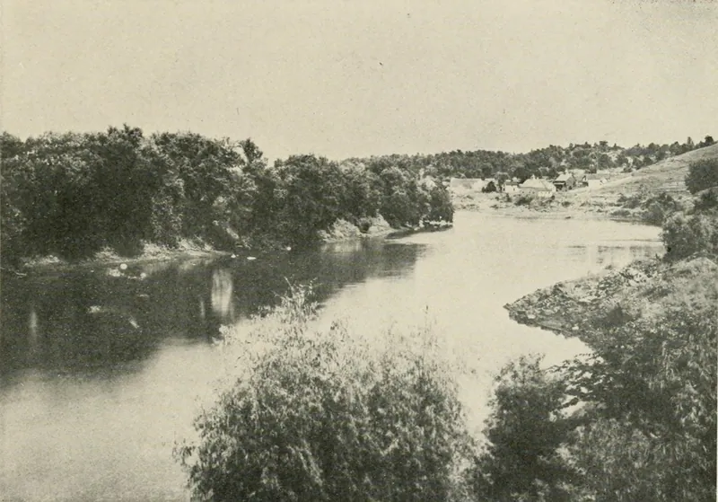 A broad river bending between wooded banks, with a cluster of small houses on an open hillside in the distance.