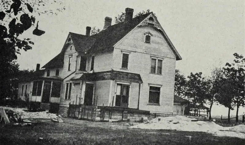 A two-story frame house with clapboard siding and three chimneys, sitting on a stone foundation with sand piles in the yard.