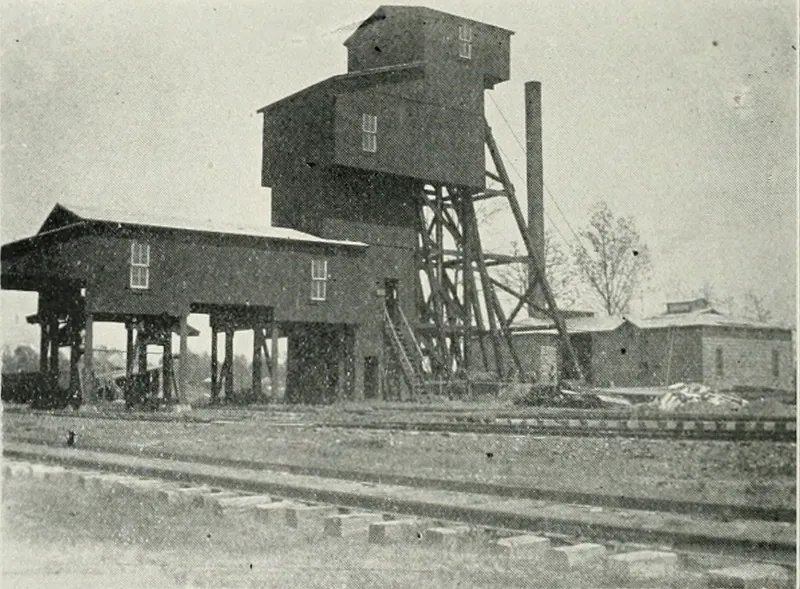 A tall wooden coal tipple and headframe with a smokestack, a long shed on wooden posts at left, and railroad tracks in the foreground.