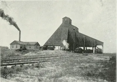 A coal mine with a large sloping wooden tipple shed, a brick boiler house with a smoking stack, and several railroad tracks across open ground.