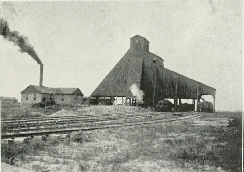A coal mine with a large sloping wooden tipple shed, a brick boiler house with a smoking stack, and several railroad tracks across open ground.