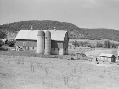 A large barn with twin silos on a hillside pasture, with a small outbuilding and a wooded ridge behind.